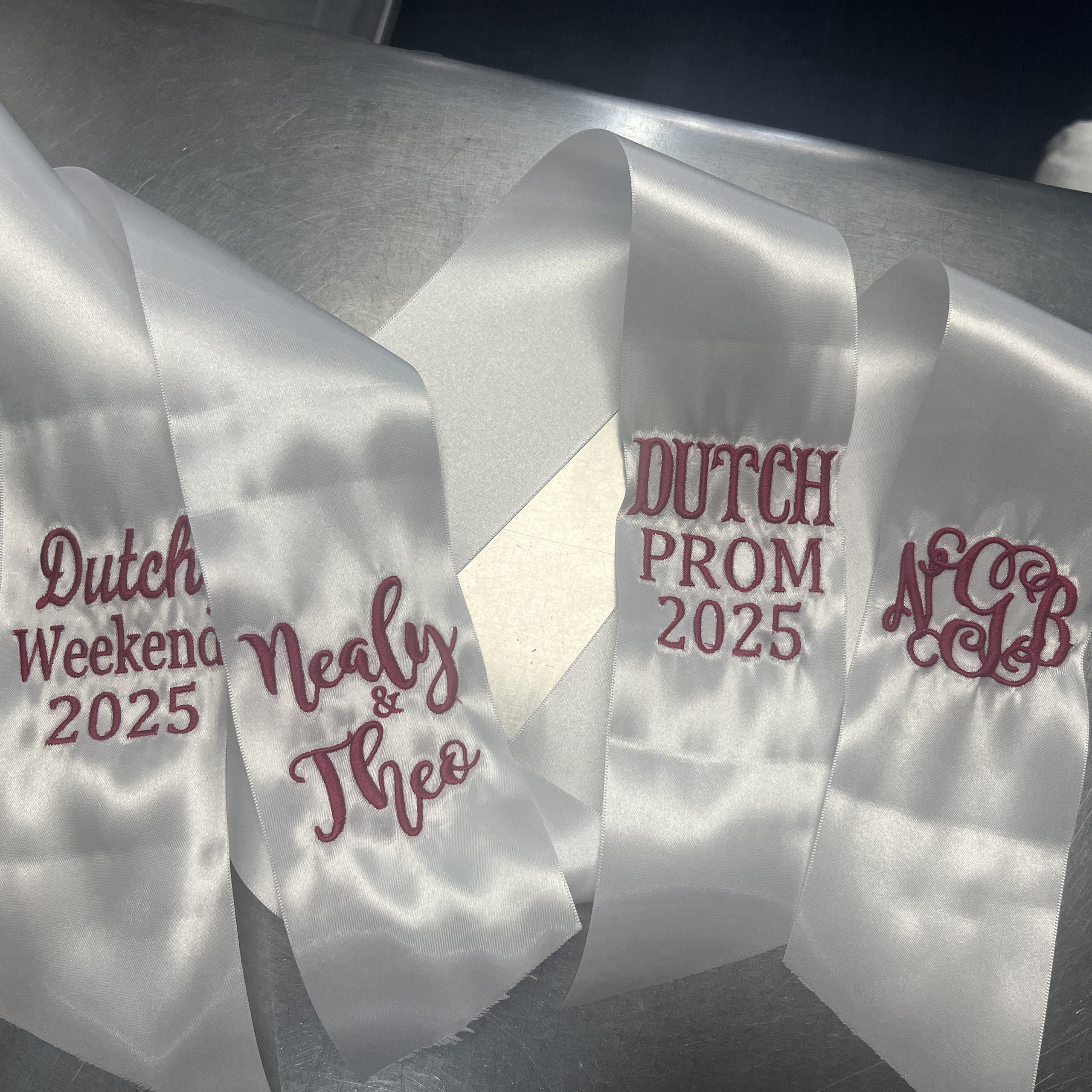 white hoco ribbons with red embroidery reading "Dutch Prom 2025" and names/monograms